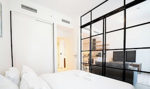 A modern hotel room with a large bed, a kitchenette, and a view through a black-framed window. - New Cozy 1BR | Dubai Hills (Dubai)
