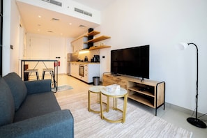 A modern living room with a sofa, a coffee table, a TV, and a kitchen in the background. - New Cozy 1BR | Dubai Hills (Dubai)