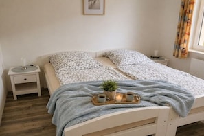 Un dormitori amb un llit, taules de nit, una tauleta solera, una bandeja amb tasses i una planta, i una finestra amb cortines.