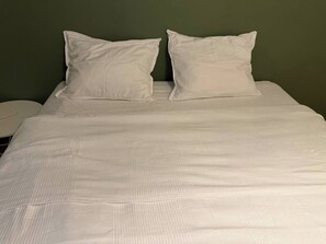 Un llit amb roba de llit i coixins blancs, una taula de nit blanca i una paret verda.