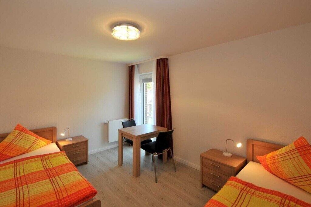 Camera d'albergo con due letti, una scrivania e una sedia. Si vede una finestra con tende, un termosifone e una luce a soffitto.
