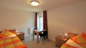 A hotel room with two beds, a desk, and a chair. There is a window with curtains, a radiator, and a ceiling light. - "Seestern", in the House of Recreation (Juist)