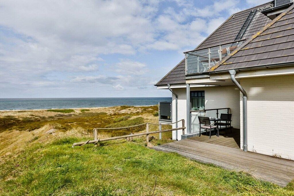 A coastal house with a deck overlooking the sea, a small outdoor seating area, and a clear view of the beach.