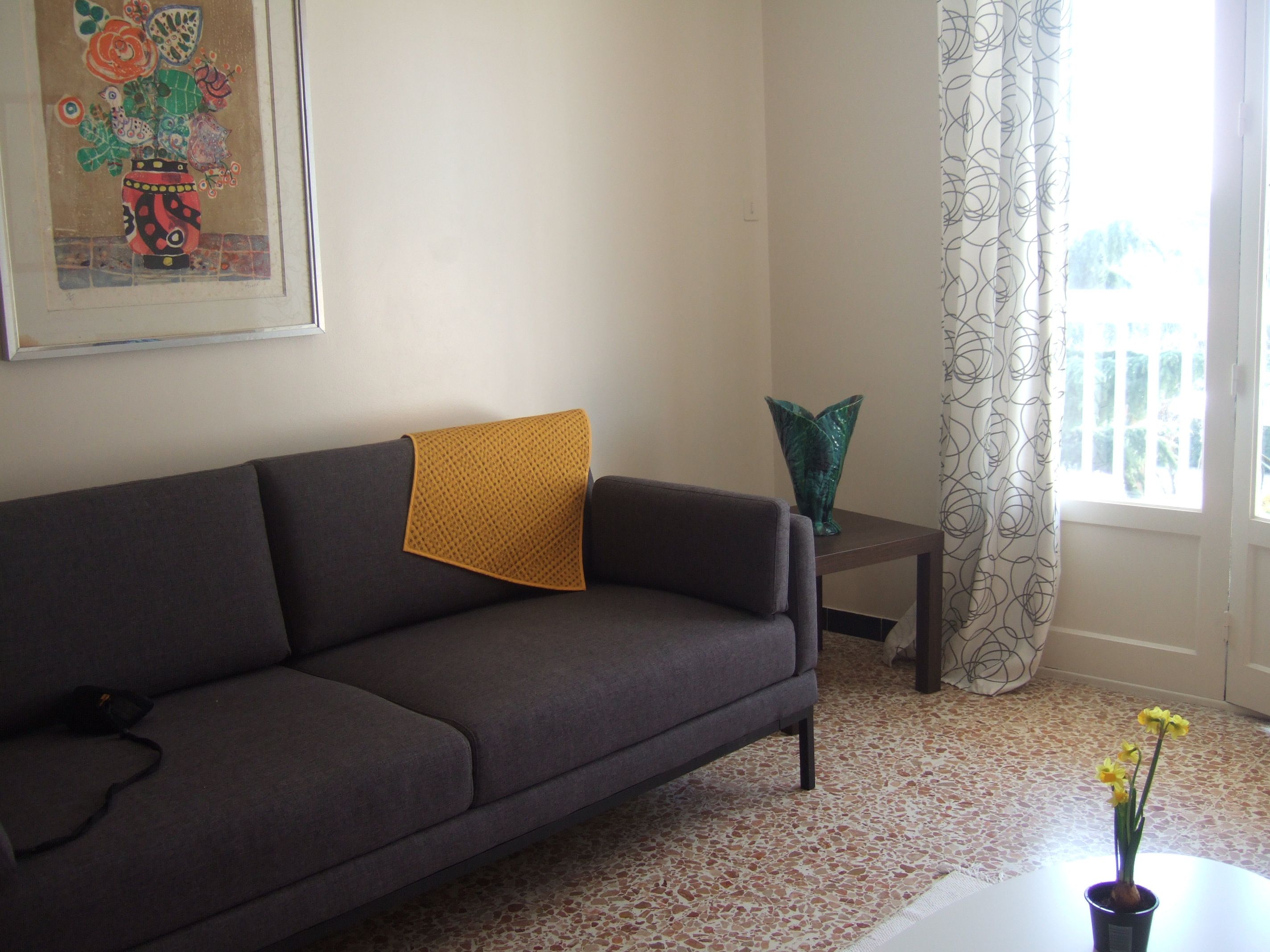 Un salon avec un canapé, une petite table sur laquelle est posé un vase de fleurs, un tableau au mur et une fenêtre avec des rideaux.