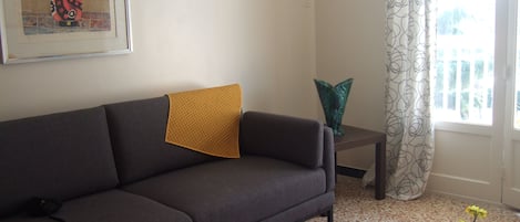 Un salon avec un canapé, une petite table sur laquelle est posé un vase de fleurs, un tableau au mur et une fenêtre avec des rideaux.