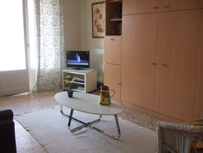 Ein Wohnzimmer mit Fernseher auf einem Schrank, einem kleinen Tisch mit Blumenvase und einem Korbstuhl.