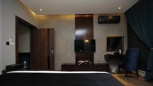 Une chambre d’hôtel avec un grand lit, un téléviseur à écran plat fixé au mur, un bureau avec une chaise et des rideaux de couleur foncée.