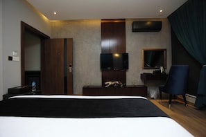 A hotel room with a large bed, a flat-screen TV mounted on the wall, a desk with a chair, and a dark-colored curtain. - Brick House Hotel  (Lahore)