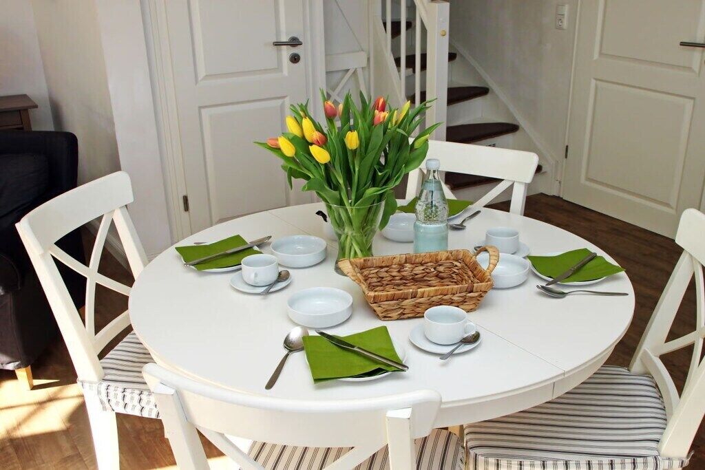 Une table de salle à manger ronde, entourée de chaises blanches, un vase de tulipes, un panier et de la vaisselle.