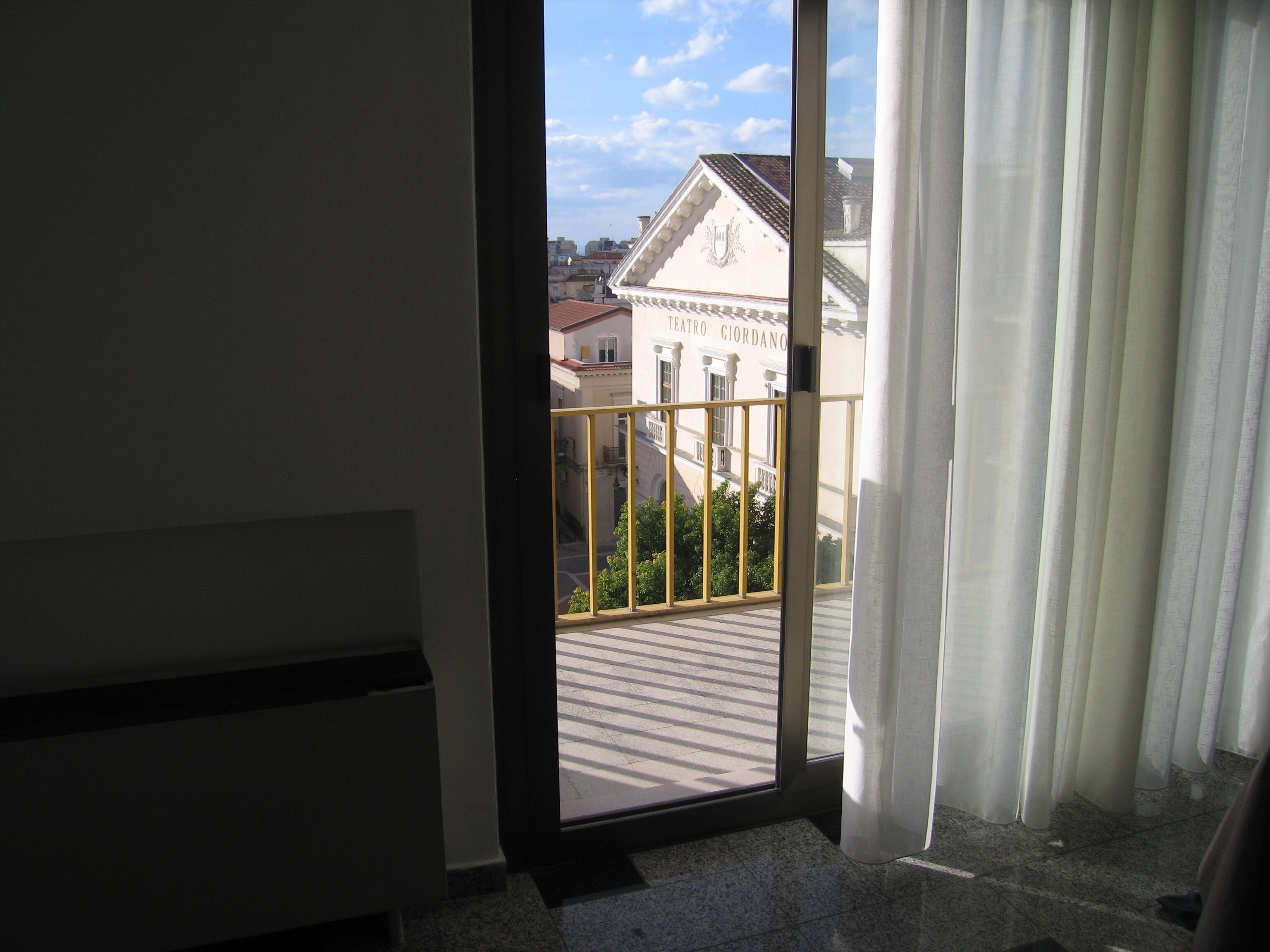 A view through a door with a balcony, a building with 'TEATRO GIORDANO' signage, and a clear sky.