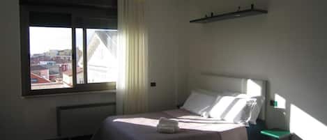 Une chambre d’hôtel avec un lit, une fenêtre avec des rideaux, une table de chevet et une vue sur les bâtiments environnants.