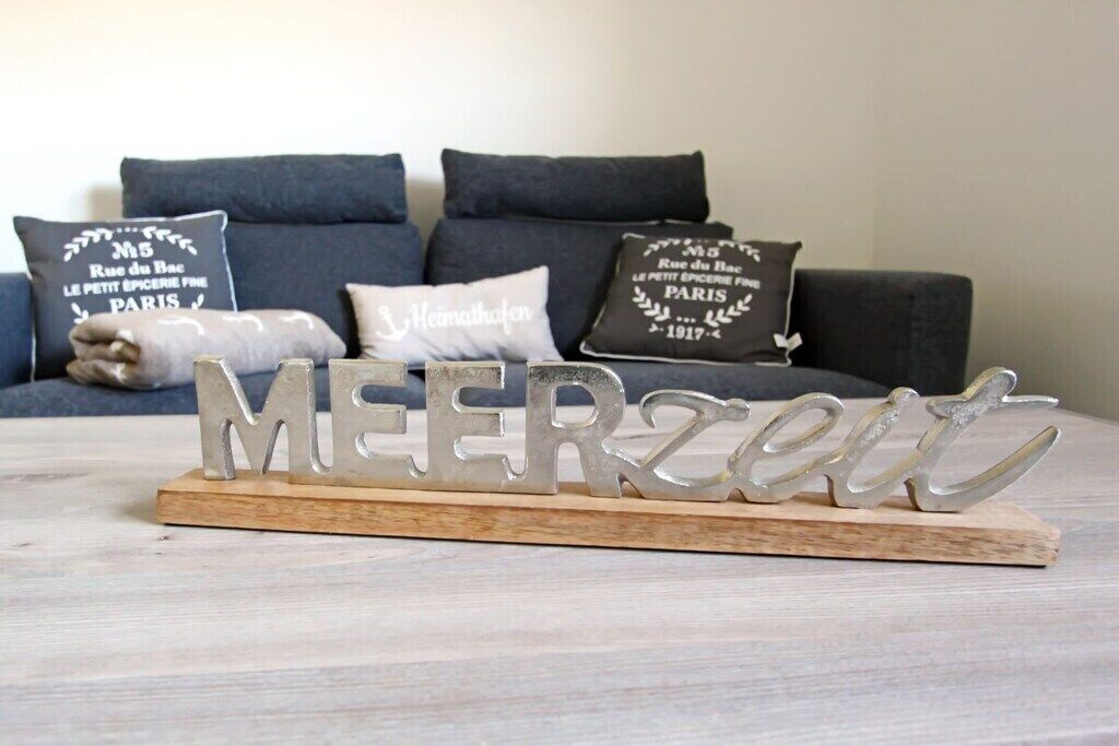 A decorative metal sign spelling 'MEERzeit' on a wooden base, placed on a light-colored wooden surface.