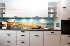 A kitchen with white cabinets, a black countertop, and a mural of a coastal landscape. - Holiday apartment with 2 bedrooms (Hörnum \/ Sylt)