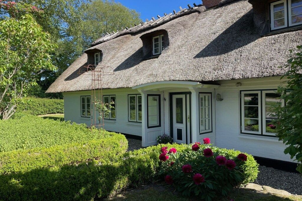 Ein traditionelles Reetdachhaus mit weißer Fassade, umgeben von gepflegten Hecken und blühenden Pflanzen.