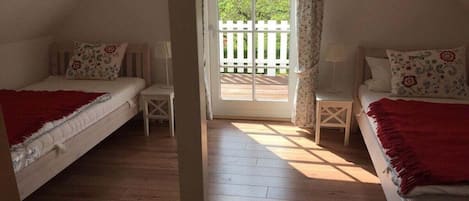 A bedroom with two beds, a wooden floor, a white door leading to an outdoor area, and a small table between the beds.