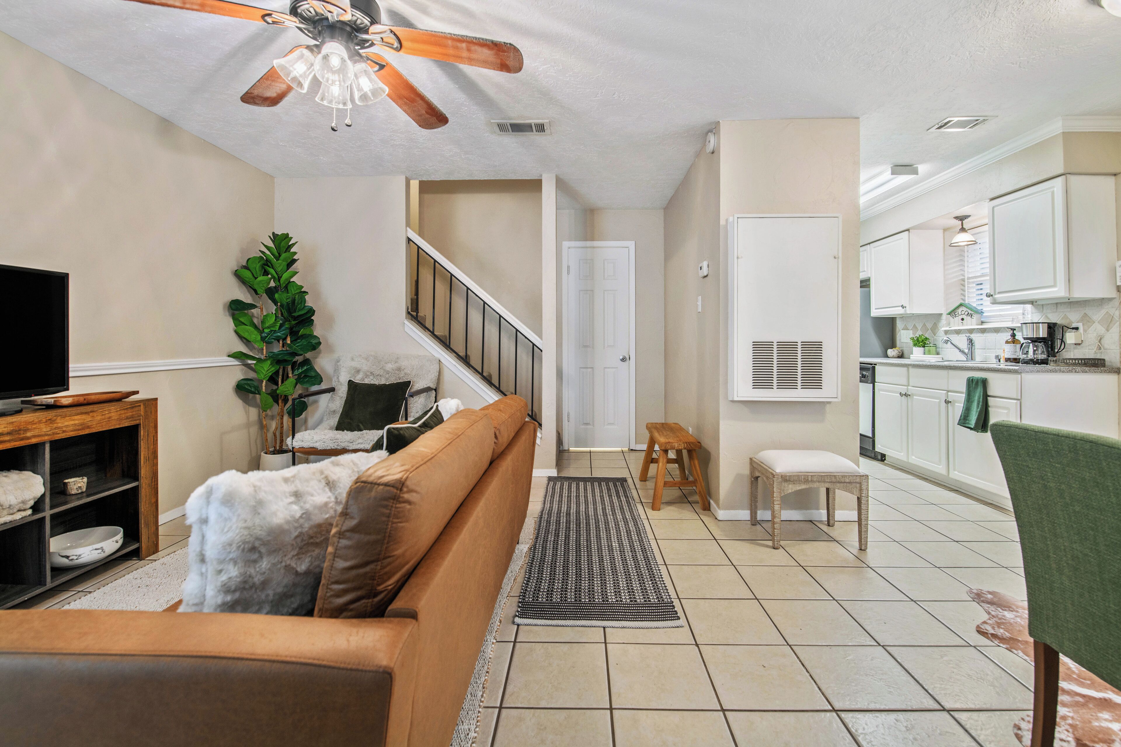 Un salon comprenant un canapé, un téléviseur, un ventilateur de plafond, un escalier et un coin cuisine avec des placards et un évier.