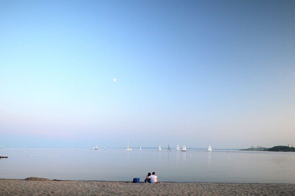 Une plage avec une mer calme, quelques voiliers et deux personnes assises sur le sable.