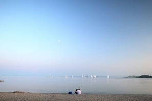 Ein Strand mit ruhiger See, einigen Segelbooten und zwei Personen, die im Sand sitzen.