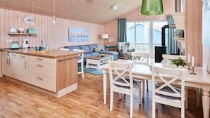 A cozy cabin interior with a wooden ceiling, a dining area with white chairs, a sofa, and a kitchen area with a wooden island. - Pilot station type G (Neustadt\/Holstein - OT Pelzerhaken)