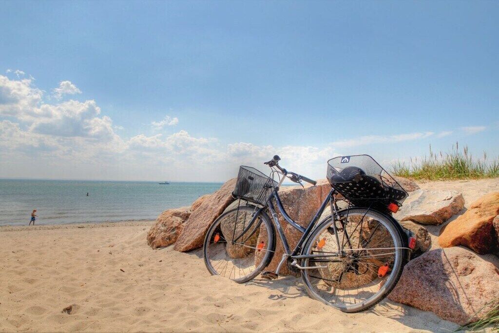A bicycle parked on a sandy beach with large rocks, a clear sky, and the ocean in the background.
