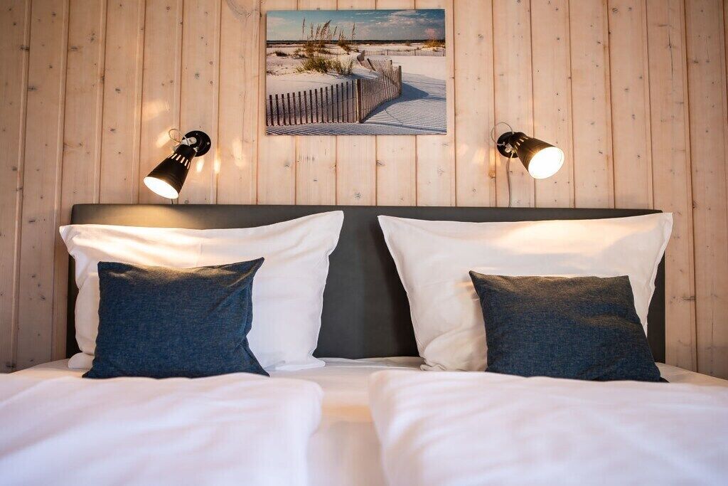 A double bed with white and dark blue pillows, two wall-mounted lamps, and a framed picture of a beach scene.