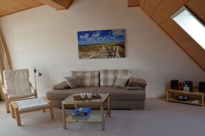 A cozy living room with a high ceiling, a sofa, a chair, a coffee table, and a painting of a beach. - Holiday apartment with 1 bedroom (Husum\/ Nordsee)