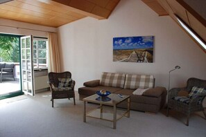 Un salon comprenant un canapé, deux fauteuils en osier, une table basse en verre, un tableau accroché au mur et une porte donnant sur un espace extérieur.