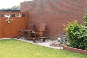 Eine hölzerne Sitzgruppe im Freien mit Tisch und Stühlen, eine Ziegelwand und ein kleiner Garten mit Wasserspiel.