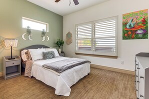 Quarto com cama, mesa de cabeceira, abajur, janela com persianas, espelho fixado na parede e um quadro.