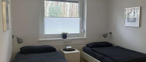 Un alojamiento pequeño, una habitación individual con dos camas individuales, una ventana con persianas, una mesita de noche con lámpara y un radiador.