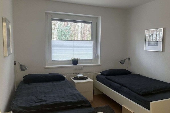 Eine kleine, einfache Unterkunft mit zwei Einzelbetten, einem Fenster mit Jalousien, einem Nachttisch mit Lampe und einer Heizung.