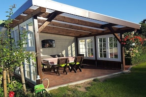 Eine überdachte Terrasse mit Tisch und Stühlen, ein Backsteinboden und Blick auf ein Haus mit großen Fenstern.