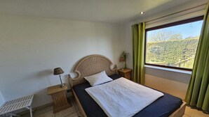 A bedroom with a bed, a nightstand, a lamp, a window with curtains, and a view of a hillside. - Baltic Sea View Schönhagen (Schönhagen)