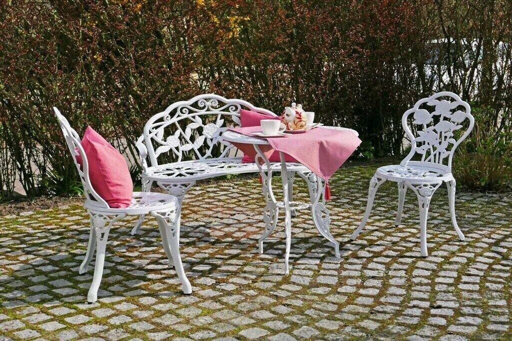 Un área de estar al aire libre con mobiliario metálico blanco, una mesa, sillas y un mantel rosa, con un fondo de follaje denso.