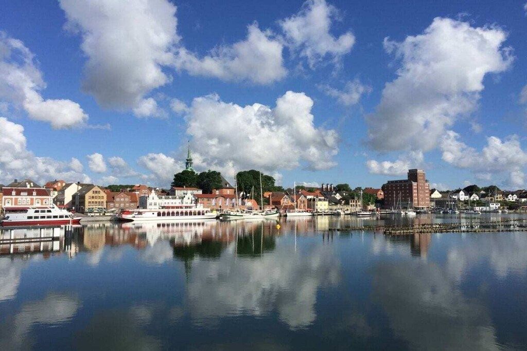 A waterfront cityscape with buildings, a dock, and a calm body of water reflecting the sky and clouds.