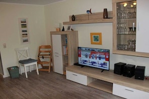 A living room with a television, wooden shelves, a chair, and a small table. - Holiday home in Dangast with swimming pool (Dangast)
