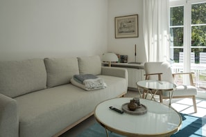 A living room with a sofa, a round glass coffee table, and a chair. There is a framed picture on the wall and a window with curtains. - Friesenring 17c - Cozy Vacation Rental at Friesenring 17c in Wenningstedt (Wenningstedt-Braderup (Sylt))