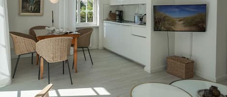 Eine moderne Küche mit weißen Schränken, einem Essbereich mit Korbstühlen, einem runden Tisch und einem an der Wand befestigten Flachbildfernseher.