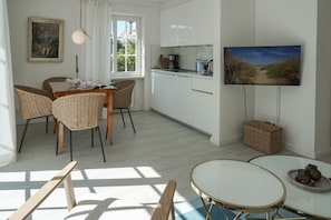 A modern kitchen with white cabinets, a dining area with wicker chairs, a round table, and a flat-screen TV mounted on the wall. - Friesenring 17c - Cozy Vacation Rental at Friesenring 17c in Wenningstedt (Wenningstedt-Braderup (Sylt))