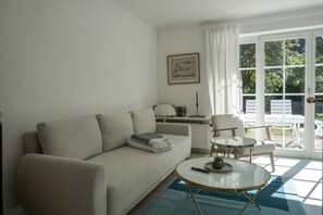 Ein Wohnzimmer mit einer Couch, einem Couchtisch und einem Sessel. Es gibt ein Fenster mit Vorhängen, eine Heizung und ein gerahmtes Bild an der Wand.