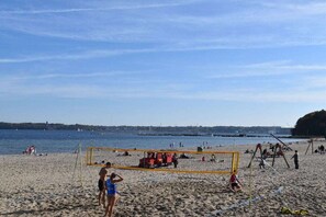 Uma praia com uma rede de voleibol, pessoas jogando e um mar calmo ao fundo.