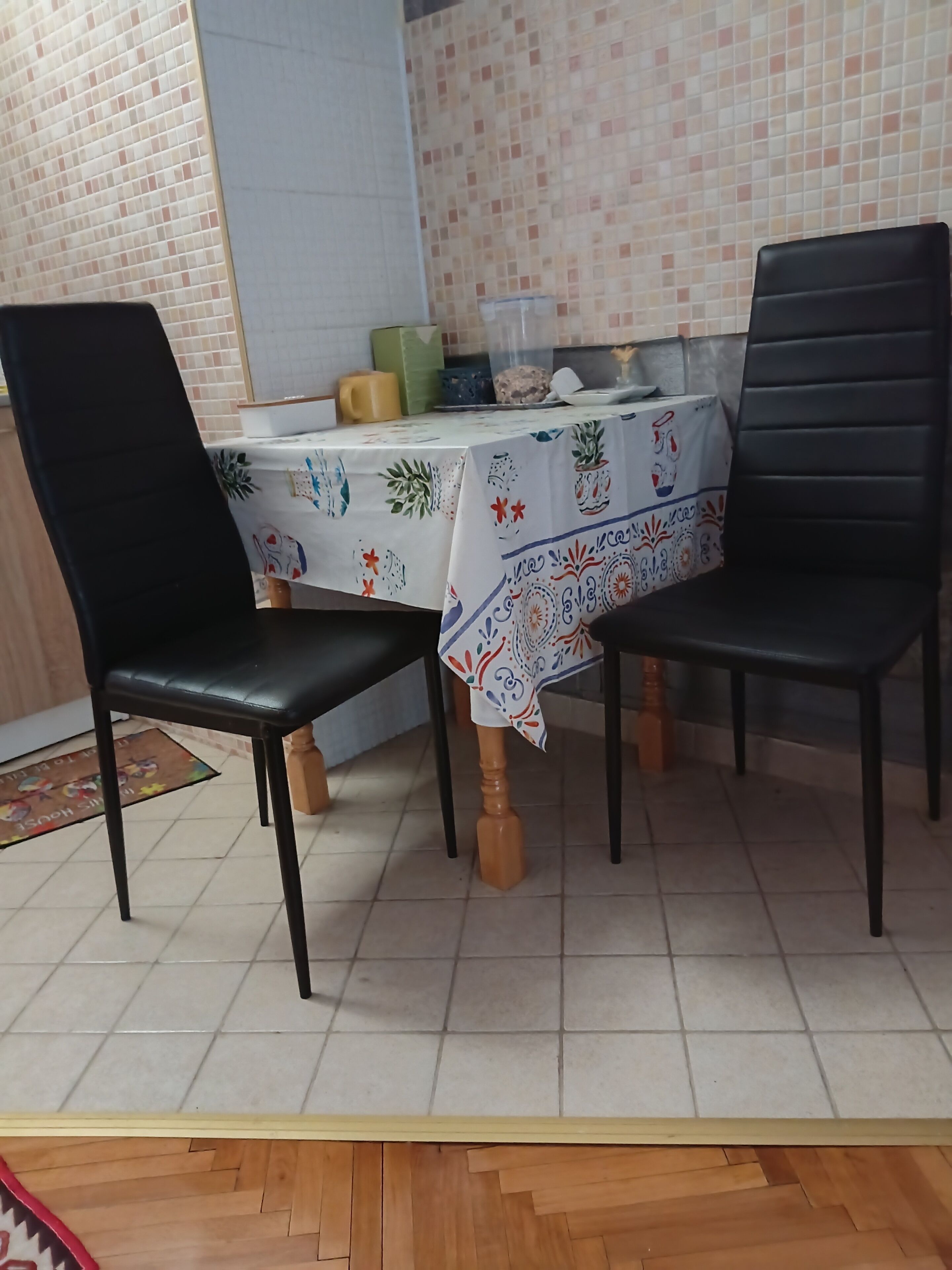 Et spiseområde med et bord dækket af en mønstret dug, to sorte stole og et flisebeklædt baggrundspanel.