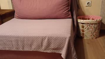 Una cama con colcha y almohadas estampadas, suelo de madera, ventana con cortinas translúcidas y un cubo de basura con diseño floral.