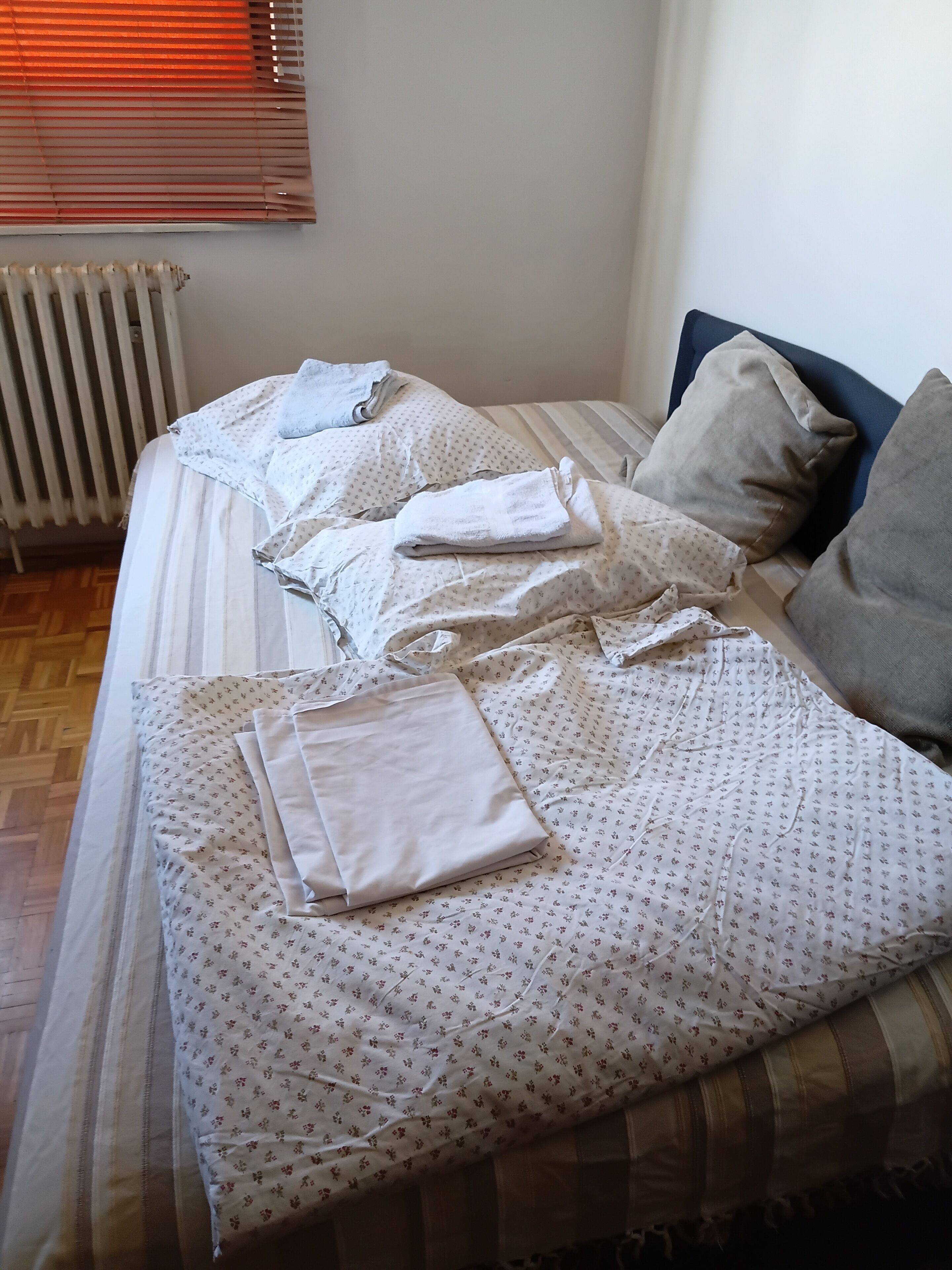 En seng med sengetæppe og puder med blomstermønster, en radiator og et vindue med persienner.