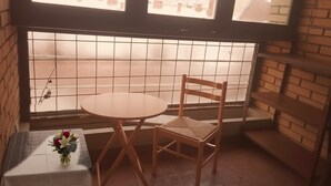 Una habitación de esquina con ventana, una mesita con un jarrón de flores y una silla de madera.