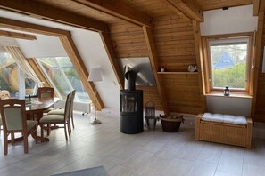 Ein gemütliches Zimmer mit Holzboden, einem Esstisch mit Stühlen, einem schwarzen Herd und einer Korbsitzbank.