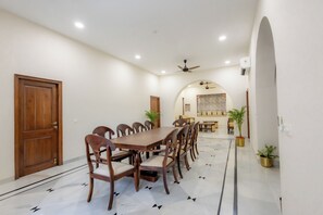 A dining area with a long wooden table and chairs, a ceiling fan, and an arched doorway leading to a living room. - Utamber House (Jodhpur)
