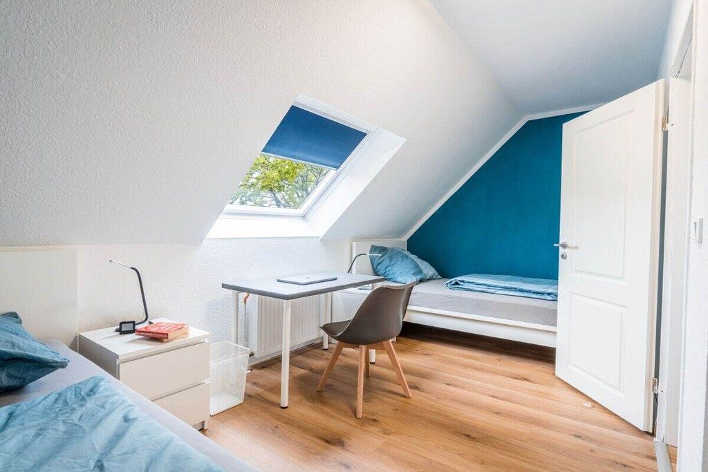 Una habitación pequeña con techo inclinado, tragaluz, cama, escritorio y silla.