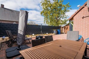 An outdoor patio area with a wooden table and chairs, a covered seating area, and a brick building in the background. - Holiday home starboard (Wittmund OT Carolinensiel)