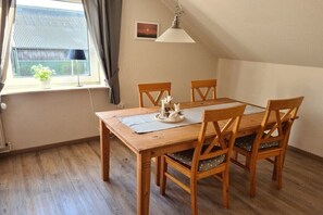 Ein Essbereich mit Holztisch und -stühlen, einer Hängelampe, einem Fenster mit Vorhängen und einer kleinen Topfpflanze.